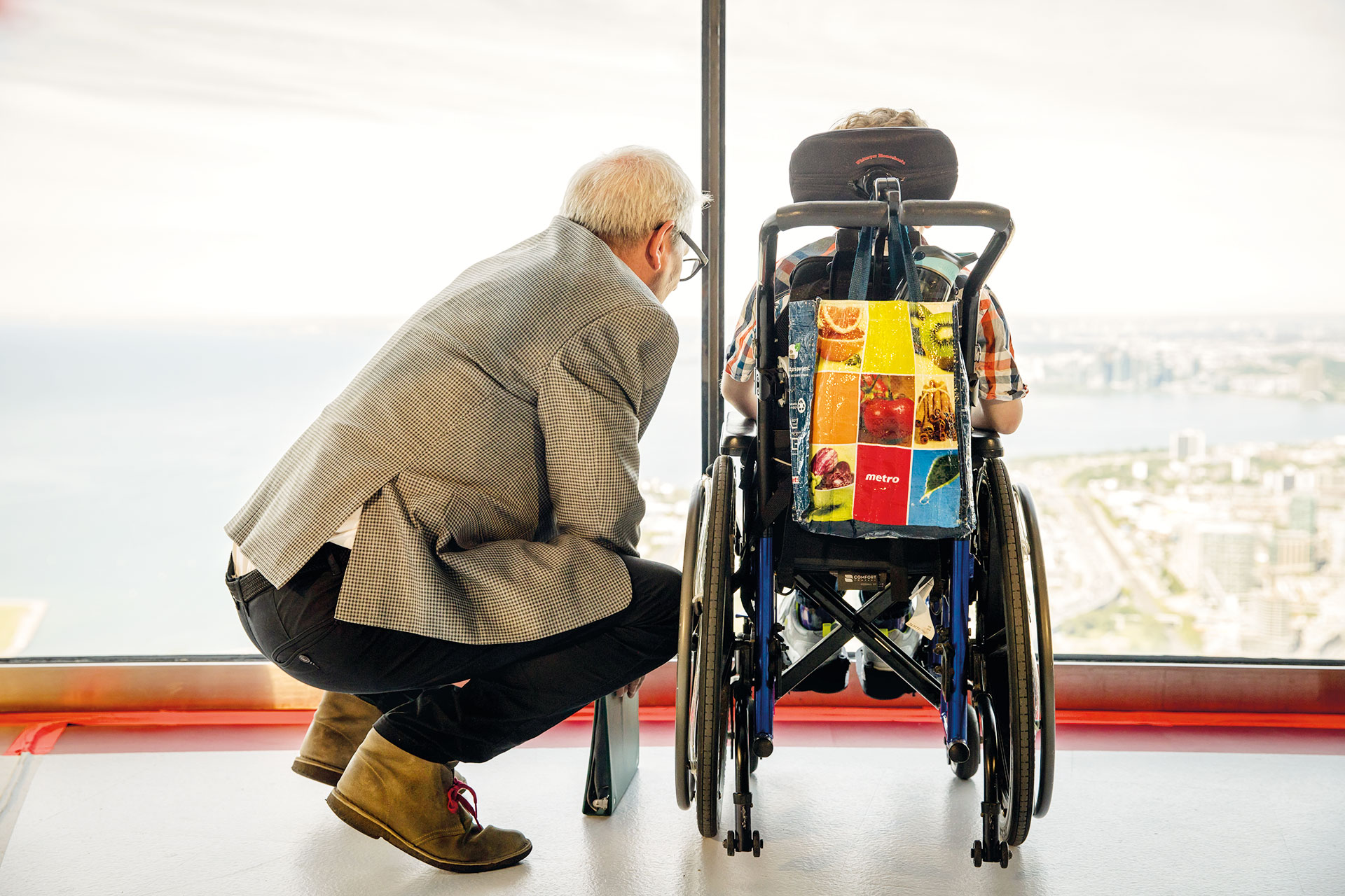 Young boy in a wheelchair and a crouching man looking out the window at the CN Tower