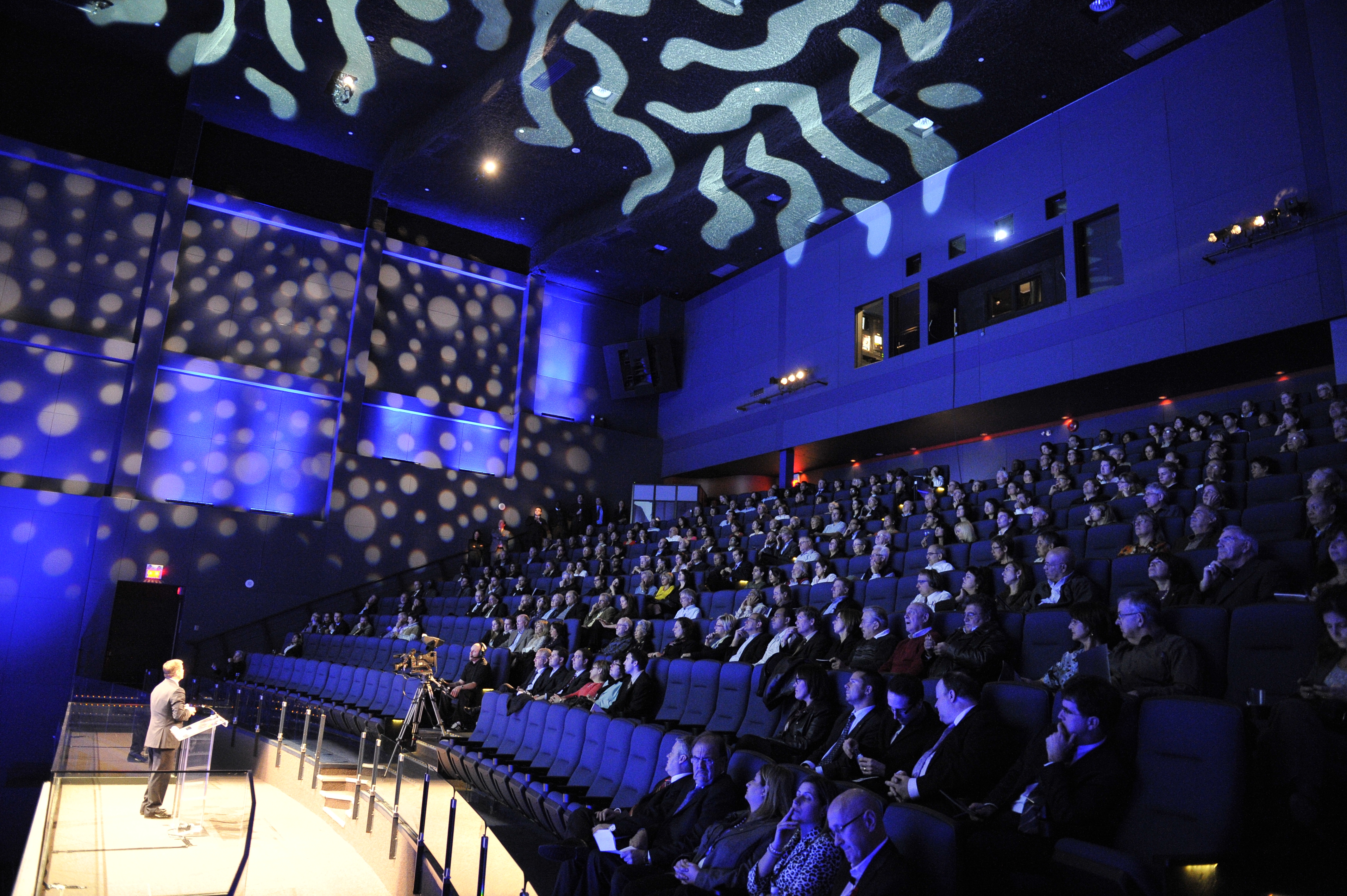 An image showing someone on stage in front of the IMAX screen in front of a large audience