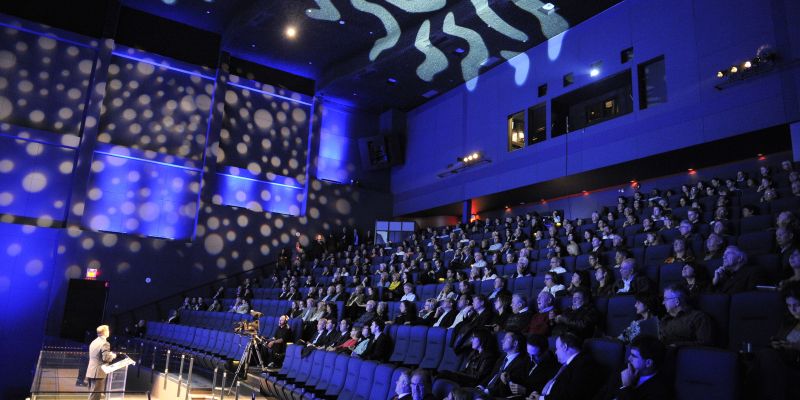 An image showing someone on stage in front of the IMAX screen in front of a large audience